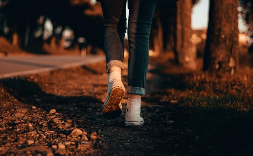A person in jeans and sneakers walks along a forest path at sunset in Italy.